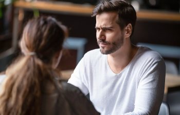 A man with a skeptical expression is sitting at a table, facing a woman with her back to the camera in a casual interior setting.