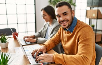 Two people working on laptops at a desk, one in a brown sweater smiling at the camera.