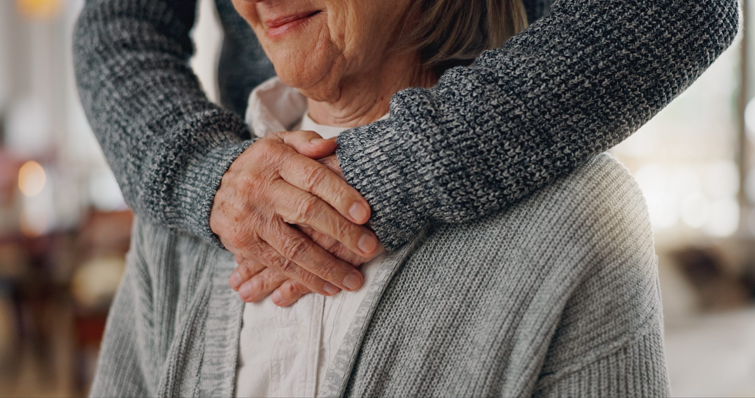 An elderly person is gently embraced from behind by someone wearing a gray knitted sweater.